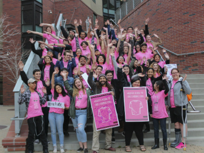  A large group of students stand on steps all wearing pink shirts with their hands raised in the air.