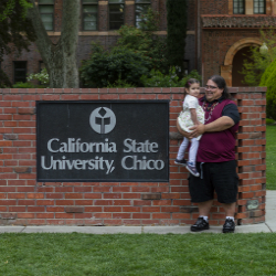 CSU, Chico student holds his young daughter in his arms in front of the CSU, Chico sign on the front lawn.